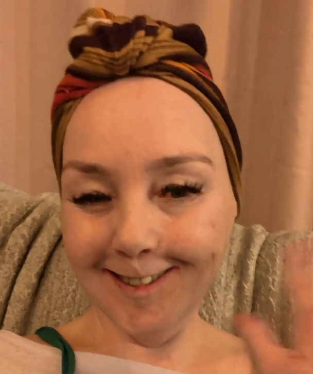 Sha, the Wheely Happy Aardvark, waves at the camera with a tired but genuine smile. She's wearing a colourful patterned head wrap and a cosy grey knit, shot in warm lamplight — the kind of lighting that says "it's late, I nearly didn't do this, but here I am anyway." 💜