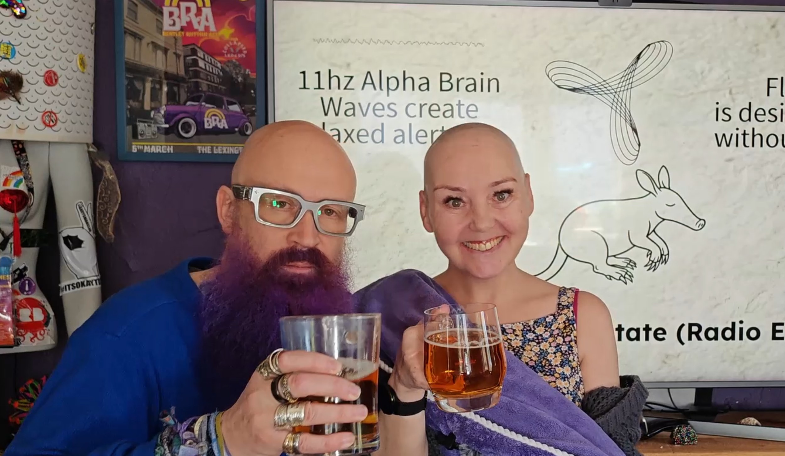 Paul (theaardvark) and Sha Taylor raise a celebratory glass together after the big shave. Paul sports his trademark purple beard, silver-framed glasses, and an impressive collection of rings, with a blue jacket and festival wristbands. Sha, freshly and gloriously bald, is wrapped in her purple towel and grinning broadly. Behind them, the AarDHD aardvark illustration is projected on the screen. Job done. 💜