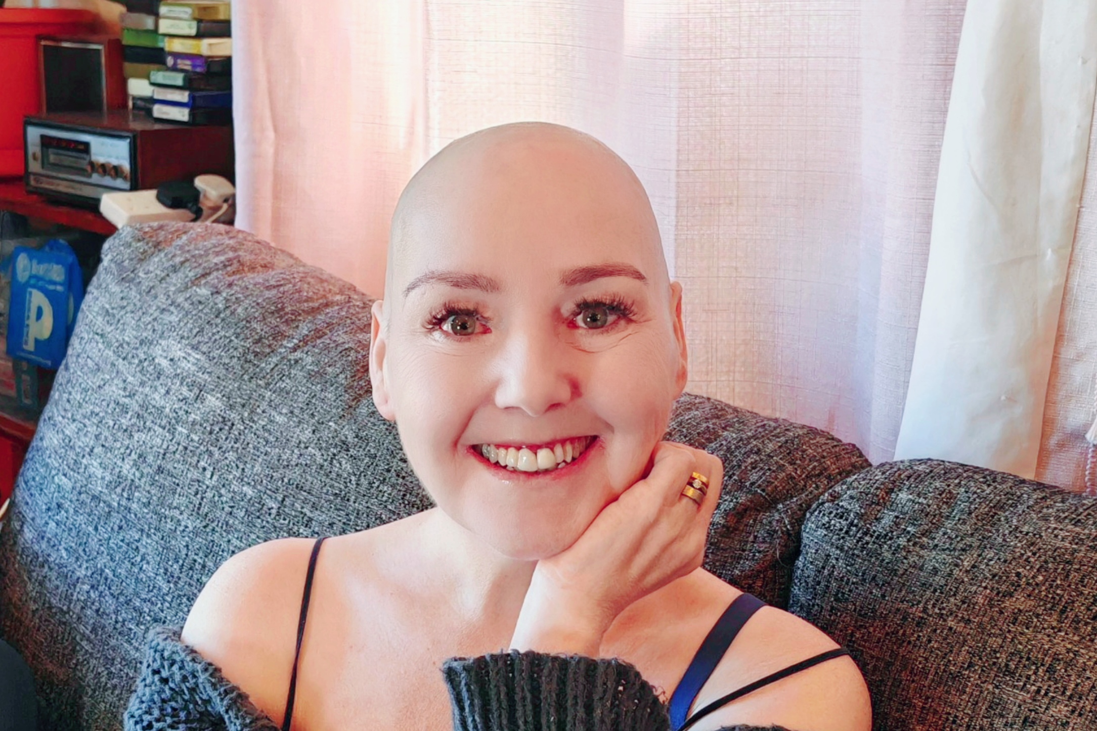 Sha Taylor — The Wheely Happy Aardvark — sitting on a grey sofa, smiling directly at the camera with her newly shaved head. She's resting her chin on her hand, showing off a gold stacked ring, and wearing a grey off-shoulder knit top over a navy cami. Her eyes are bright and her smile is wide. She looks completely at ease and, honestly, absolutely radiant. 💜