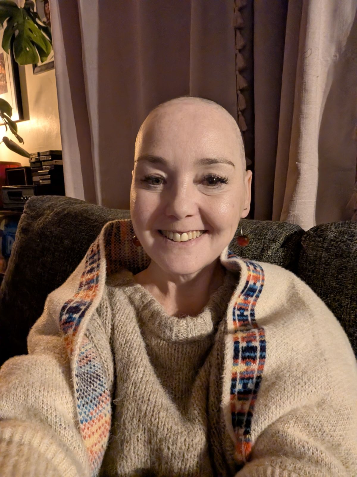 Sha back at home in the evening, settled into a soft armchair. She has a shaved head, a gentle smile, earrings in, and is wrapped in a cream knitted cardigan with a colourful patterned trim. The room is warmly lit and lived-in. a plant, a lamp, curtains drawn against the dark. She looks tired but at peace.