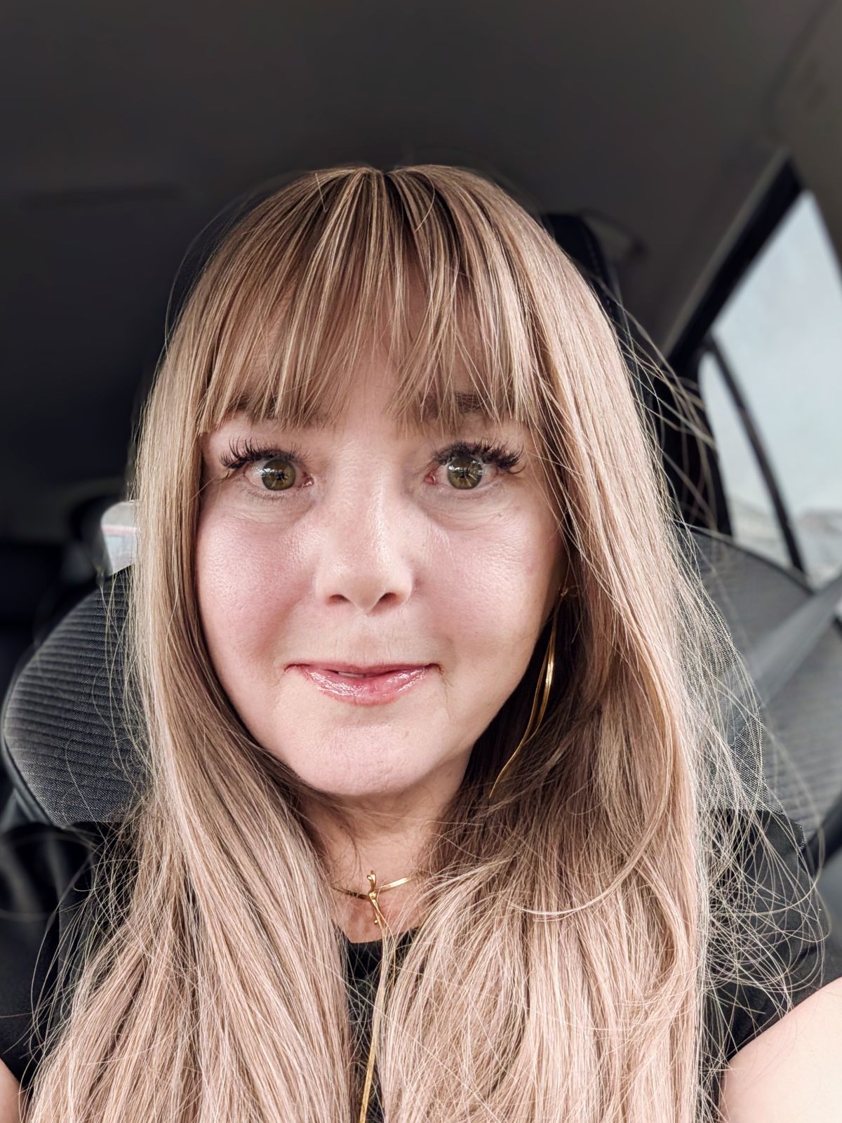 Sha sits in the passenger seat of a car, ready for the funeral. She wears a long honey-blonde wig with a fringe, gold hoop earrings, a delicate gold necklace and a black top. Her expression is soft and composed. Calm on the outside, nervous underneath.