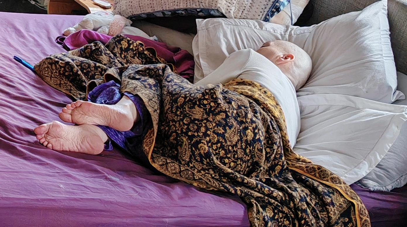 Sha lies curled on her side on a purple bed, wrapped in a large dark paisley blanket, bare feet poking out of the bottom. Her shaved head rests on a white pillow. She is asleep. The overall impression is of someone who has absolutely, completely run out of steam.
