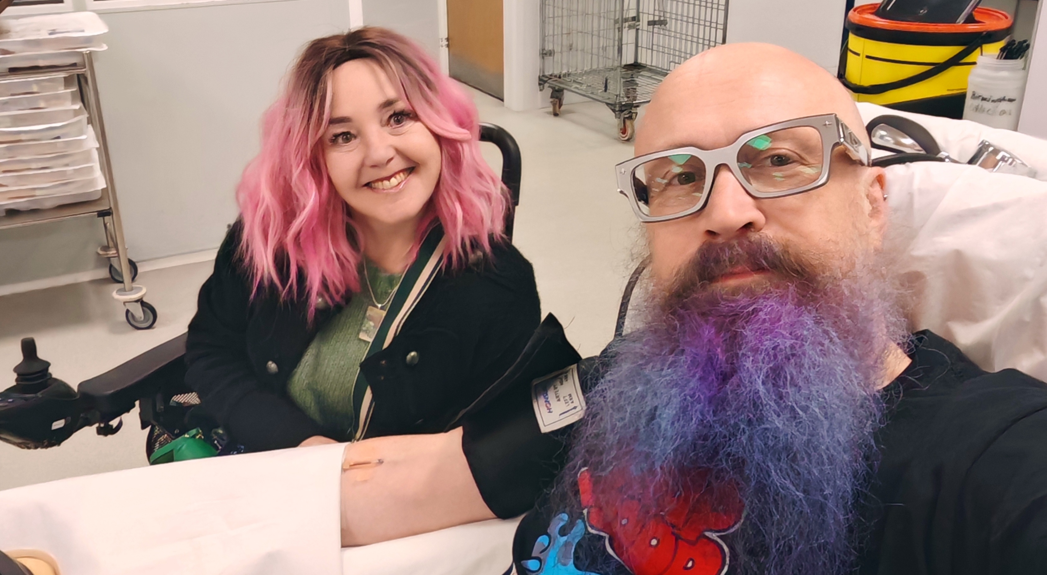 Selfie of Paul and MrsVark at an NHS platelet donation centre. Paul is reclining in a donor chair in the foreground, bald with a vivid purple beard, grey thick-framed glasses, and a black graphic t-shirt featuring cartoon characters. A donation needle is visible in his arm. MrsVark sits alongside him, smiling, with pink wavy hair and a black jacket over a green top. Medical equipment and storage units are visible in the background.
