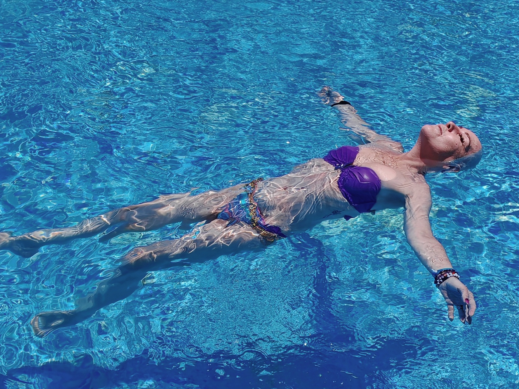 A woman in a purple bikini floating on her back in a vivid blue swimming pool, arms outstretched, eyes closed and face tilted up towards the sun. She has short white hair and colourful beaded bracelets on her wrists. Her body is fully relaxed, suspended in the clear water.
