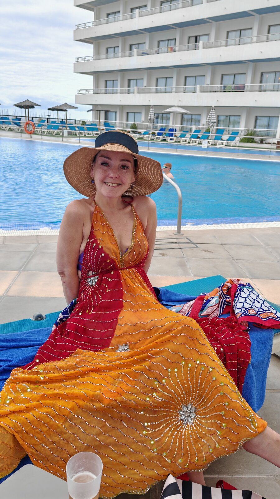 Sha sitting poolside at the hotel, wearing a wide-brimmed sun hat and an orange, red and gold sequinned maxi dress, smiling at the camera. Sun loungers and hotel balconies behind her.