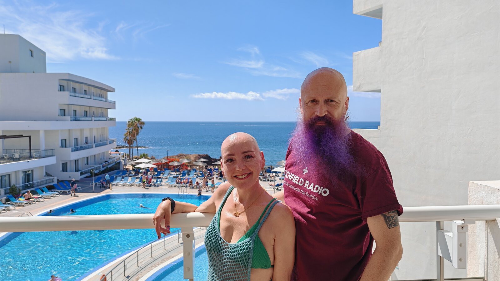 Sha and Paul standing together on a hotel balcony overlooking the pool and sea. Sha wears a teal vest top, Paul wears a maroon Lichfield Radio t-shirt with his beard dyed purple.