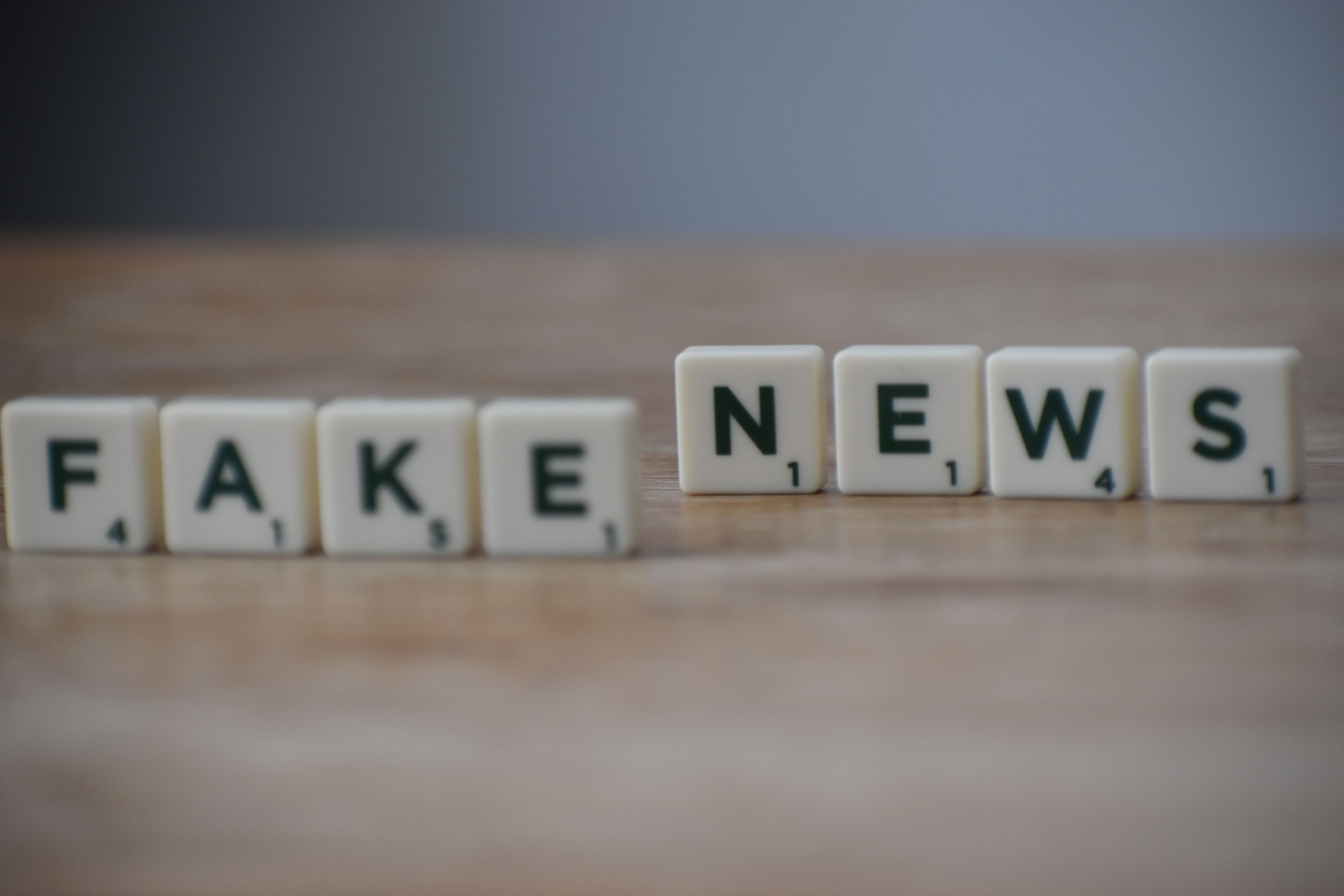 Scrabble tiles spelling "FAKE NEWS" arranged in two groups — “FAKE” slightly blurred on left, “NEWS” sharply focused on right — rest on a warm-toned wooden surface against a muted cobalt blue backdrop; the composition uses shallow depth of field to draw attention to the word “NEWS,” visually emphasizing its current prominence and urgency amid misinformation.