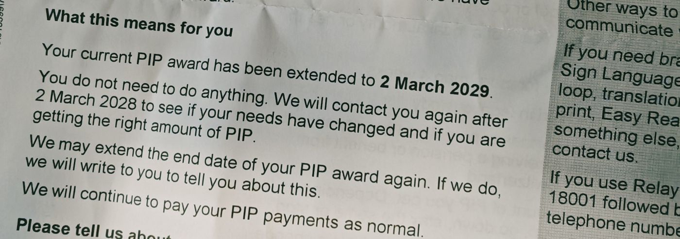 document: A close-up shot of a government notice explaining that a PIP (Personal Independence Payment) award has been extended to 2 March 2029 — no action needed from the recipient. The text reassures the reader that payments will continue normally, and future reviews are scheduled for 2 March 2028 with possible further extensions. Visible on the right is a partial view of accessibility options like sign language loop, Easy Read print, and Relay telephone service contact details. The paper appears slightly crumpled with soft shadows suggesting indoor lighting; it’s clear from the tone that this document brings relief and continuity to its recipient.