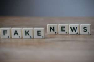 Scrabble tiles spelling "FAKE NEWS" arranged in two groups — “FAKE” slightly blurred on left, “NEWS” sharply focused on right — rest on a warm-toned wooden surface against a muted cobalt blue backdrop; the composition uses shallow depth of field to draw attention to the word “NEWS,” visually emphasizing its current prominence and urgency amid misinformation.