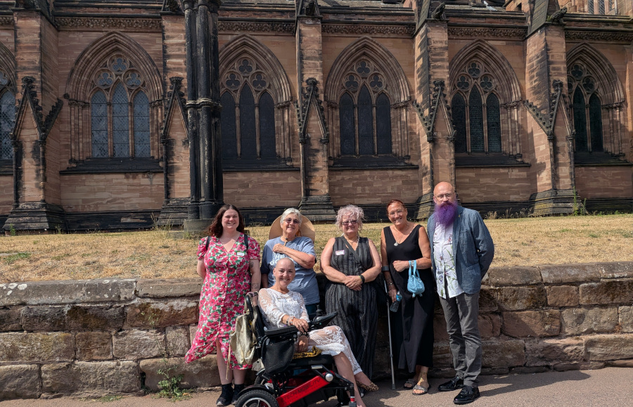 group: Six members of the Burntwood Spoonies pose together on a sunny day in front of Lichfield Cathedral’s ornate Gothic facade, featuring pointed arches and dark stone detailing against warm sandstone walls. From left to right: a woman in a vibrant pink floral dress smiles beside a woman with short grey hair wearing a wide-brimmed hat; next is MrsVark, seated in a black powered wheelchair with red accents, smiling warmly while holding a beige bag; beside her, an older woman with silver-grey hair and glasses rests her hands over her lap; then another woman in a black sleeveless dress holds a small blue handbag; finally, on the far right, “theaardvark”, bald with a long, vivid purple beard, wears a light-blue jacket over a white patterned shirt and grey trousers. The group stands against a low stone wall with dry grass behind them. All are smiling — clearly happy to be together in this picturesque setting under bright sunlight.