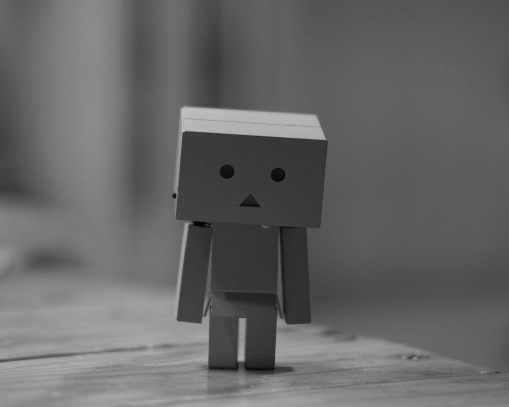 figurine: A monochrome, softly lit shot of a small, boxy Danbo-style figurine standing on a wooden surface. Its simple, geometric form — square head, rectangular body, stubby limbs — is rendered in muted greys, with two black dot eyes and a triangular mouth suggesting a neutral or contemplative expression. The shallow depth of field blurs the background into gentle, indistinct tones, focusing all attention on the little figure’s quiet presence.